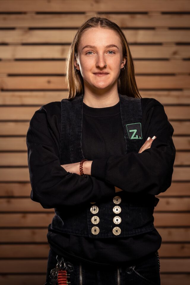 Frau in schwarzer Arbeitskleidung vor Holzlattenwand, Zimmerei Freiburg.