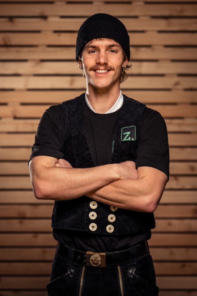 Mann mit Mütze vor Holzpaneelen, lächelnd, mit Armkreuz.