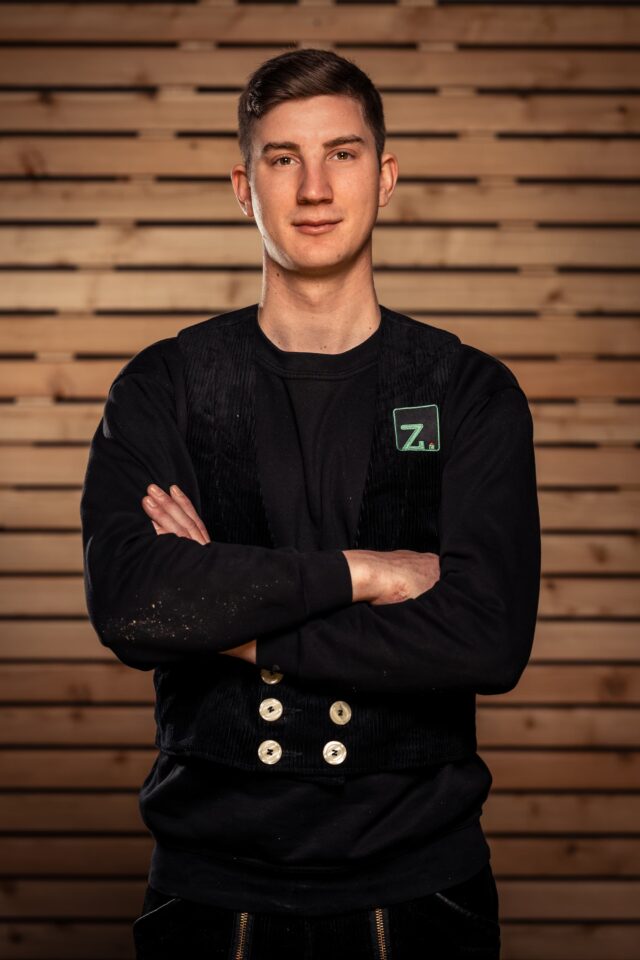 Junger Mann mit schwarzen Jacke vor Holzvertäfelung, Zimmerei Freiburg.